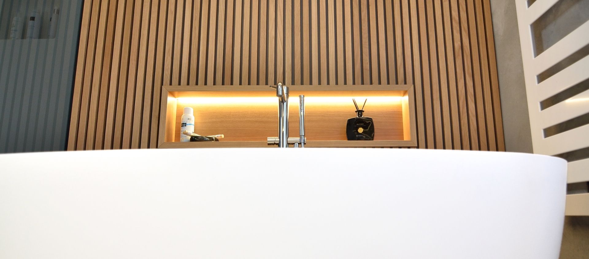Weiße freistehende Badewanne vor einer Wand mit vertikalen Holzlatten und einem beleuchteten Regal mit Dekoartikeln